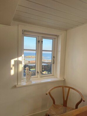 Una habitación con un ventanal blanco que da a la playa, una silla de madera y una estatua blanca en el alféizar.