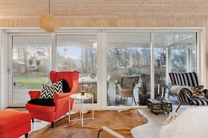 A modern living room with a large sliding glass door, a red armchair, a striped armchair, and a white fur rug. - Cosy private cottage 800 m from the beach (Løkken)