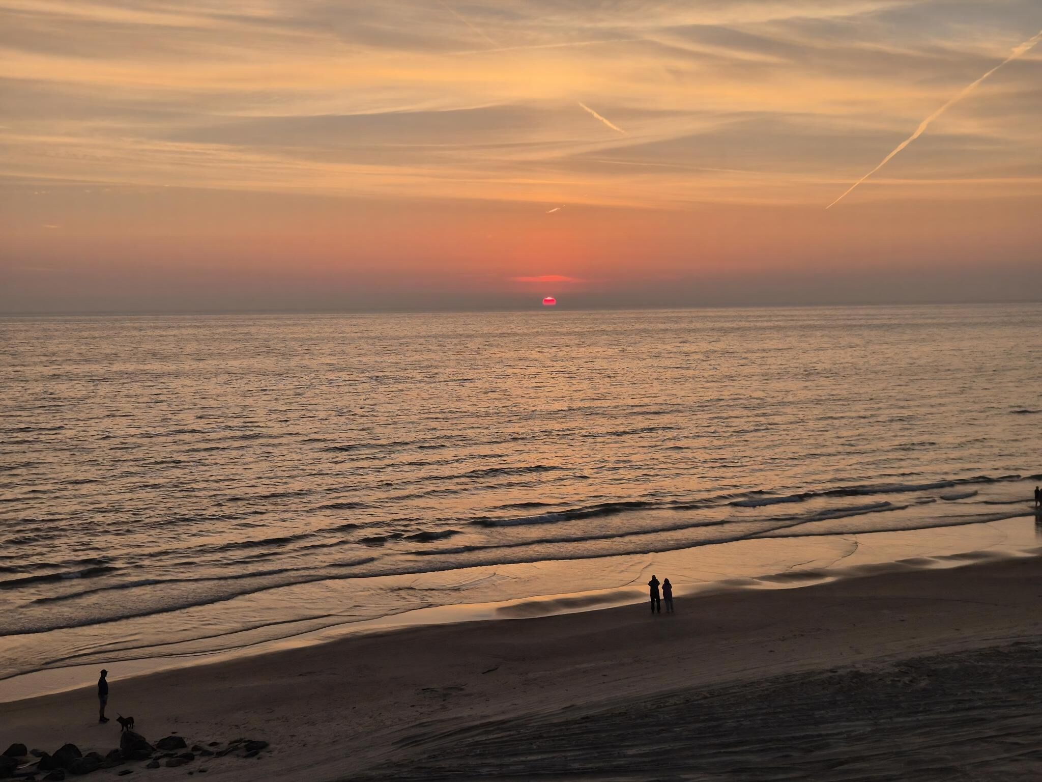 Een strand bij zonsondergang, met golven en enkele mensen die lopen.