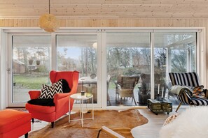 A modern living room with a large sliding glass door, a red armchair, a striped armchair, and a white fur rug. - Cosy private cottage 800 m from the beach (Løkken)