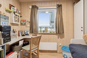 A room with a wooden interior, a window with curtains, a desk with various items, a chair, and a bed with a blue container. - Cosy holiday home near Legoland (Hovborg)