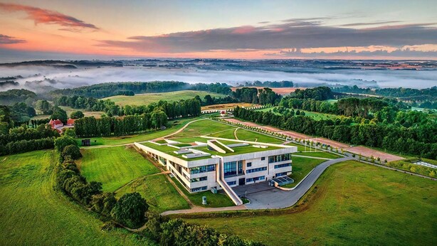 Un edificio moderno con techo plano, rodeado de campos verdes y árboles, con vistas al paisaje de fondo.