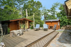 Eine Holzterrasse mit zwei Liegestühlen, einem kleinen Tisch und einer Holzhütte inmitten von Bäumen.