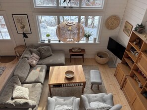 Ein modernes Wohnzimmer mit einer Couch, Sesseln, einem Couchtisch aus Holz, einem Fernseher und einem großen Fenster mit Blick auf eine verschneite Landschaft.