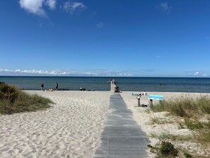 Ein sandiger Strand mit einem Betonweg zum Wasser, einige Personen und ein klarer, blauer Himmel.