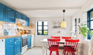 A kitchen with blue cabinets, a white countertop, a stove, and a dining area with a table and red chairs. - Cozy farmhouse close to the sea (Nakskov)