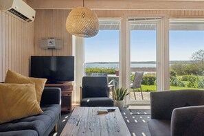 A living room with a TV, sofa, and coffee table, a potted plant, and a view of a body of water through a glass door. - Holiday home with lovely view of Lillebælt (Fredericia)