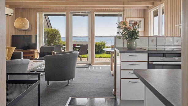 Ett modernt kök med utsikt över havet, en matplats med bord och stolar samt ett vardagsrum med soffa och TV.