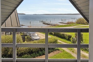 Blick durch ein Fenster mit Metallrahmen auf ein Gewässer, eine Anlegestelle, ein Auto und viel Grün.