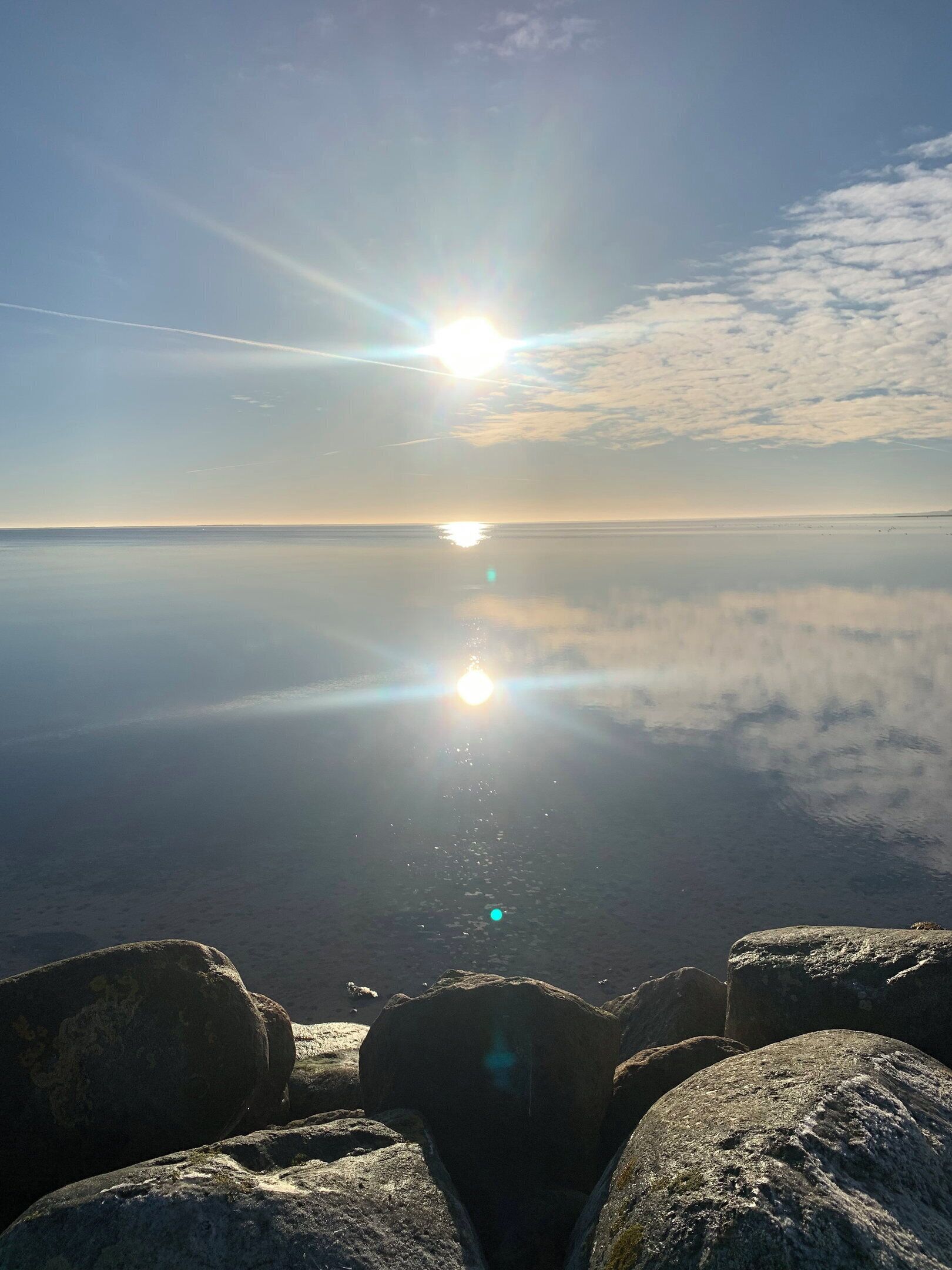 A calm body of water with a clear sky, a sun reflecting on the surface, and large rocks in the foreground.