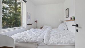 Una camera da letto con una grande finestra, un letto con biancheria bianca, un comodino e un piccolo ripiano con degli oggetti.
