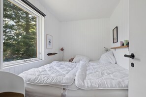 Ein Schlafzimmer mit großem Fenster, einem Bett mit weißer Bettwäsche, einem Nachttisch und einem kleinen Regal mit Gegenständen.