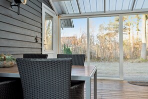 Een overdekt terras met een tafel en stoelen, een bloempot en uitzicht op bomen en gras.