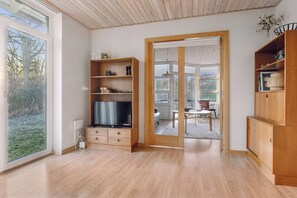 A modern interior with wooden flooring, built-in shelving, a television, and a sliding glass door leading to an outdoor area. - Cozy wooden cottage close to nature and beach (Vig)