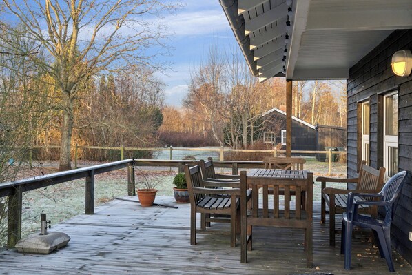 Una terraza de madera con mobiliario exterior, barandilla y vistas a árboles y un edificio al fondo.
