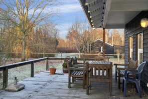 A wooden deck with outdoor furniture, a railing, and a view of trees and a building in the background. - Cozy wooden cottage close to nature and beach (Vig)