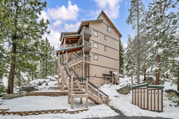 A multi-story building with a wooden staircase leading to a balcony, surrounded by snow-covered trees and rocks.