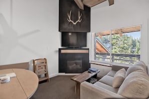 A modern living room with a fireplace, a sofa, a dining table, and a view of greenery outside.