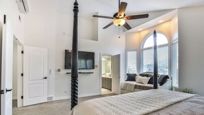 A bedroom with a large bed, a ceiling fan, a TV, and a view of the city through large windows. - Grand Mountain Retreat: Hot Tub, Sleeps 21, Sofa Sleeper on Main Floor (Draper)