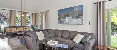 Een moderne woonkamer met een hoekbank, een salontafel, een plafondventilator en een groot raam met gordijnen.