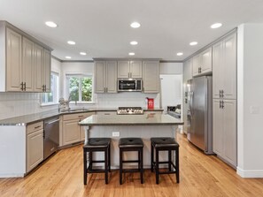 A modern kitchen with white cabinets, a central island with stools, stainless steel appliances, and wooden flooring. - Grand Mountain Retreat: Hot Tub, Sleeps 21, Fire Pit, Mountain Views (Draper)