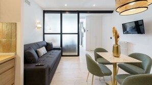 Un salon moderne avec un canapé foncé, une table à manger avec des chaises, un téléviseur fixé au mur et une grande fenêtre dotée de panneaux en verre dépoli.
