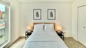 A hotel room with a large bed, two bedside tables with lamps, a window with a view, and two framed pictures on the wall. - Downtown Condo near Vancouver Skytrain (Surrey)