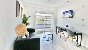A modern living room with a black sofa, a glass coffee table, and a dining area with a black table and two chairs. There is a large window with a view of buildings outside. - Downtown Condo near Vancouver Skytrain (Surrey)