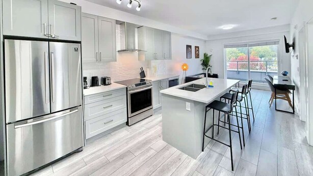 A modern kitchen with stainless steel appliances, a central island with bar stools, and a view to the outside. - Downtown Condo near Vancouver Skytrain (Surrey)