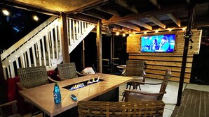 An outdoor patio with a wooden table, wicker chairs, a TV mounted on a wooden wall, and a staircase. - 5 Bdrm Beach House-Walk to Beach-Pet Friendly (North Myrtle Beach)