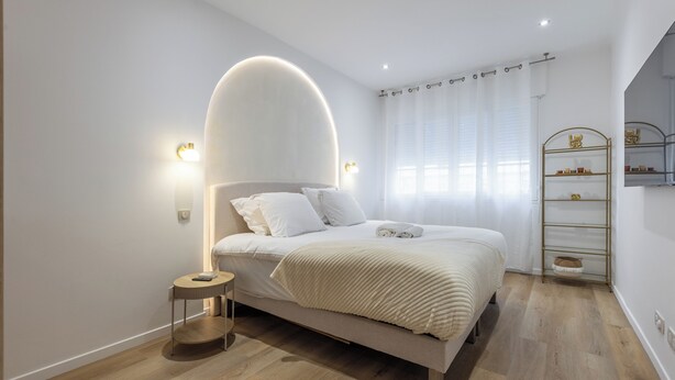 Une chambre à coucher moderne avec un lit, des lampes de chevet, une petite table ronde et une étagère contenant des articles de toilette.