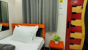 A hotel room with a bed, a nightstand with a plant, a red and yellow door, and a window with curtains.