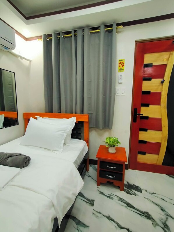 A hotel room with a bed, a nightstand with a plant, a red and yellow door, and a window with curtains. - Kabayan Hotel (Aliaga)