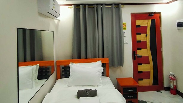 A hotel room with a bed, a bedside table, a mirror, a ceiling light fixture, a door, and an air conditioning unit. - Kabayan Hotel (Aliaga)