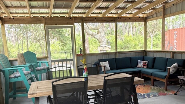A covered outdoor seating area with a wooden table and chairs, a green sofa, and a view of trees through large windows. - Sandy Paws RV Retreat...campsite, full hookups, 30-A, 4-minute drive to the Cape (Port St. Joe)