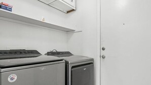 A laundry room with two washing machines, a white door, and a shelf above the machines. - Quaint 3-bedroom house with AC in charming Spring (Spring)