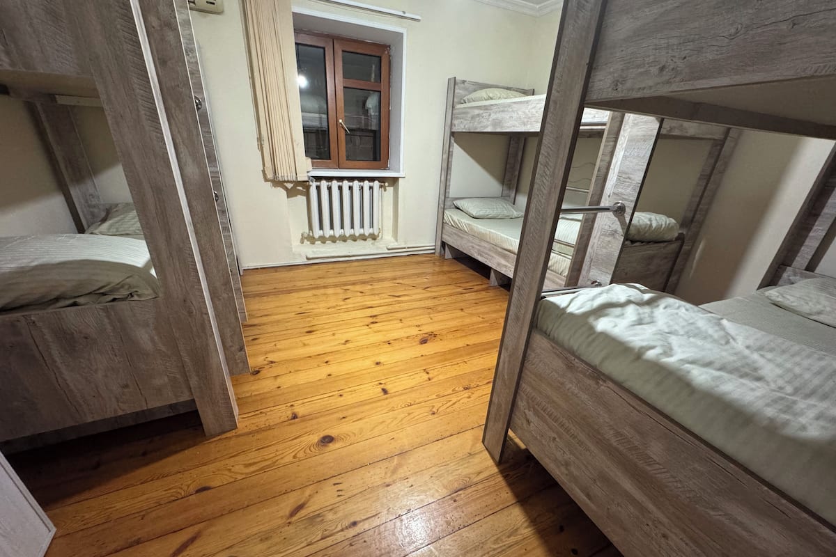 Um quarto em dormitório com beliches, soalho de madeira, uma janela com cortinas e um radiador.