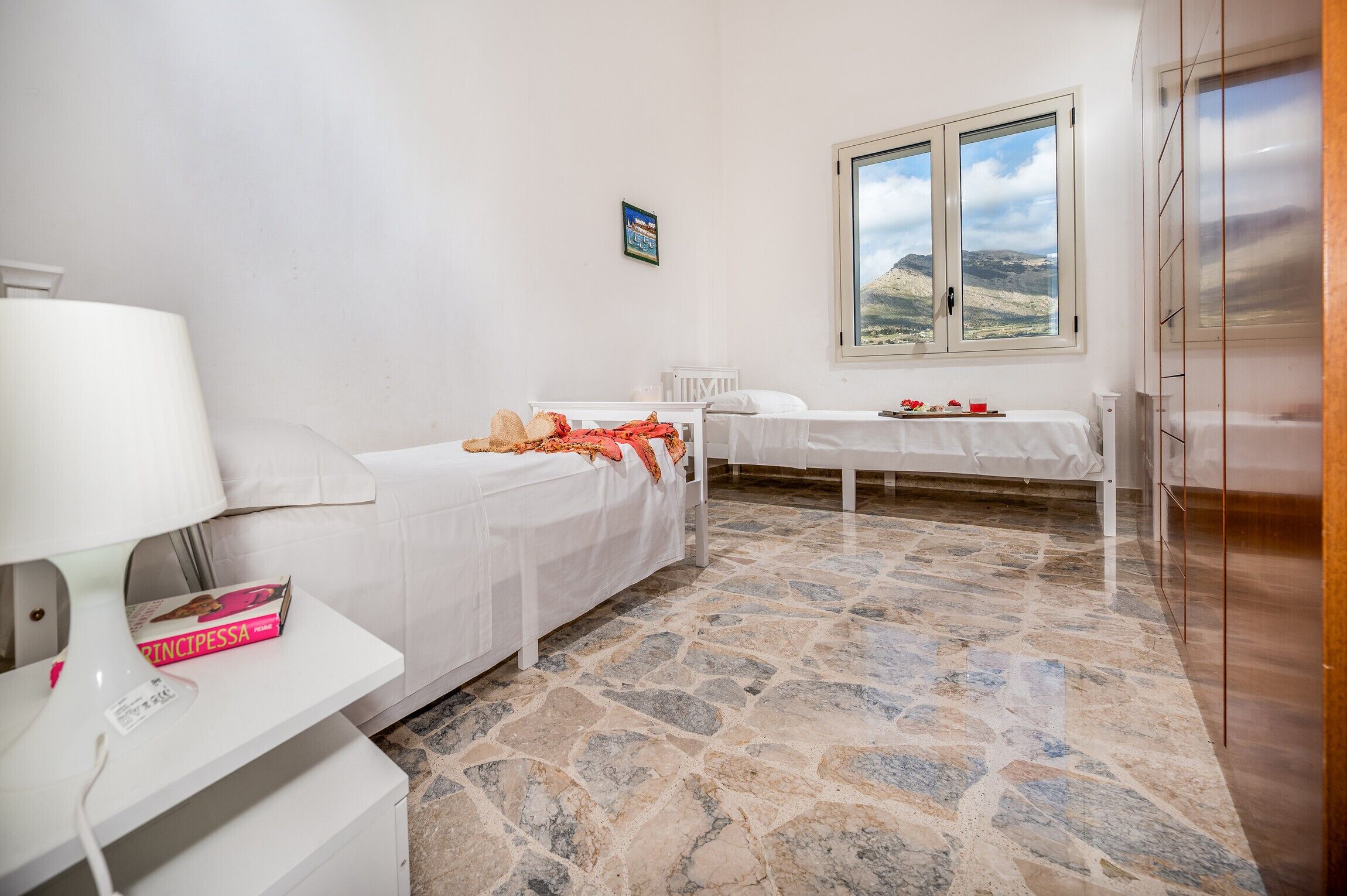 A bedroom with two beds, a stone-tiled floor, a window with a mountain view, and a white wardrobe.