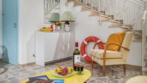 A living room with a staircase, a white cabinet with decorative items, a yellow table with fruits and a bottle, and a chair with a cushion. - Villa Le Terrazze Blu (Castellammare del Golfo)