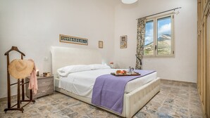 A bedroom with a bed, a nightstand, a hanging basket, a window with a view, and a wall with framed pictures. - Villa Le Terrazze Blu (Castellammare del Golfo)