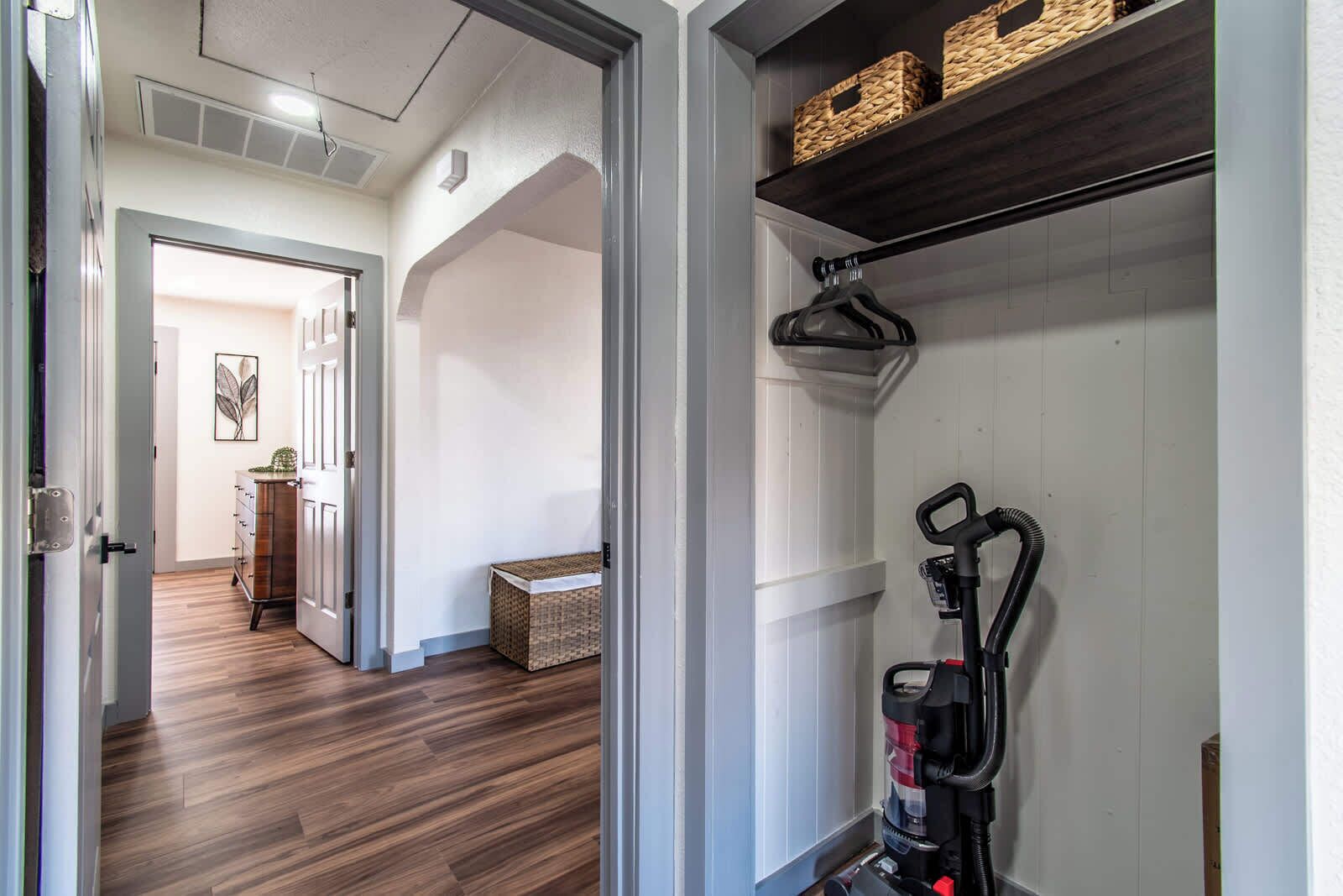A hallway with a wooden floor, a coat rack, and a vacuum cleaner. There is a door leading to another room and a bench with baskets.