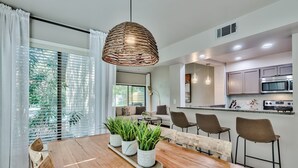 Une cuisine moderne équipée d’appareils électroménagers en acier inoxydable, un coin repas avec une table et des chaises en bois, et une grande fenêtre avec des stores.