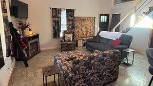 A living room with a patterned sofa, a chair with a deer pillow, a television mounted on the wall, and a staircase leading to an upper level.