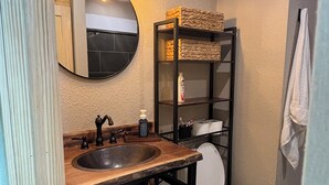 A bathroom with a wooden vanity, a round mirror, a toilet, and a shelf with baskets.