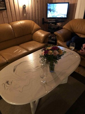 Uma sala de estar com sofá de couro marrom, uma mesa de centro branca com um vaso de flores e uma televisão ao fundo.