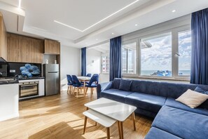 A modern kitchen with wooden cabinets, a dining area with blue chairs, a blue sofa, and a view of the sea through large windows. - Infinity Beachfront Suites by PS (Rrashbull)