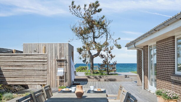 Eine Holzdar Terrasse mit Tisch und Stühlen, ein großer Baum und Blick auf das Meer.