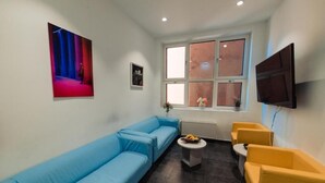 Een woonkamer met een blauwe bank, een gele fauteuil, een klein rond tafeltje met fruit, een flatscreen televisie aan de muur en een raam met uitzicht op buiten.