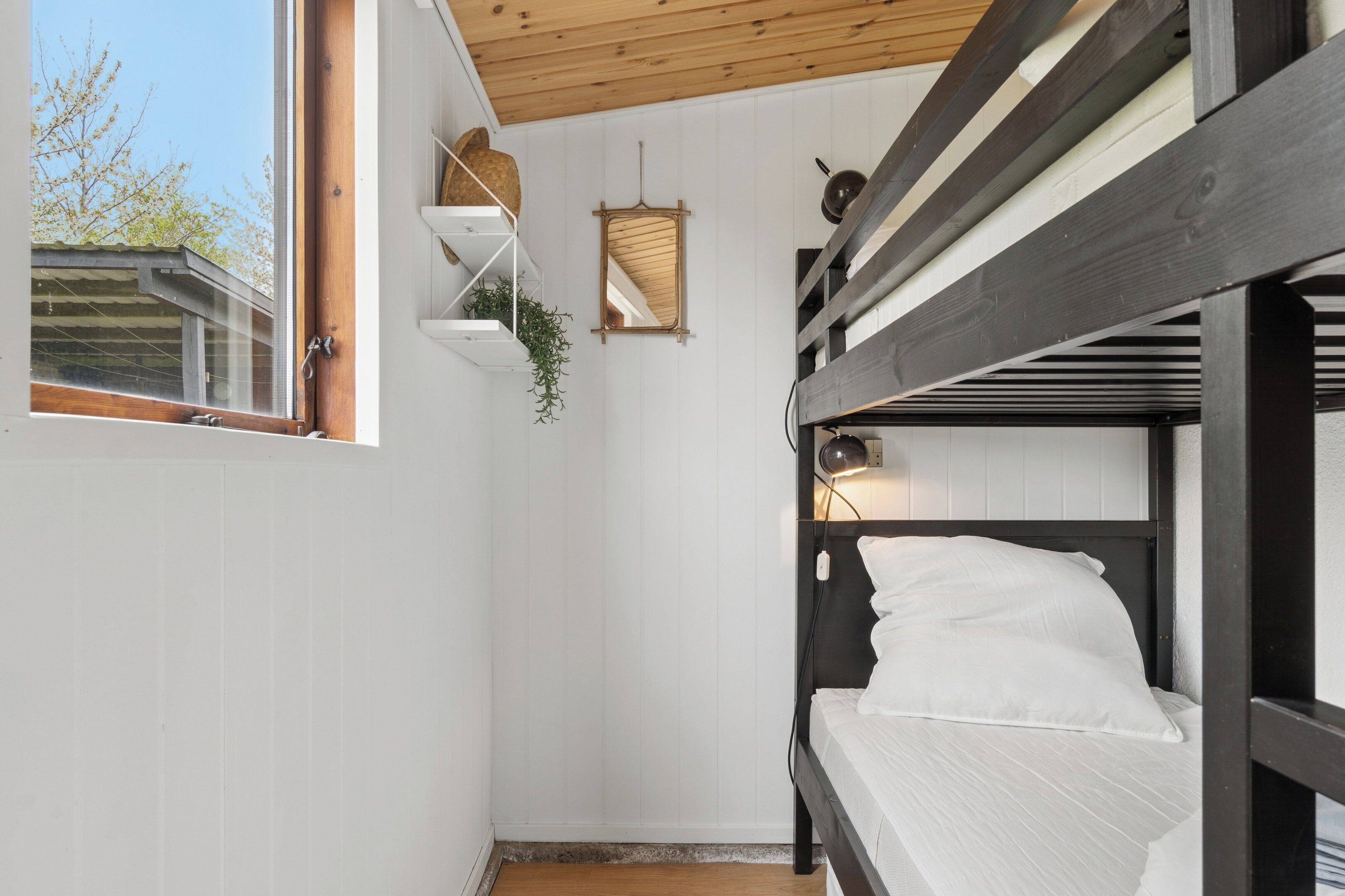 Een slaapkamer met stapelbedden, witte muren, een houten plafond, een raam en een plank met een plant.