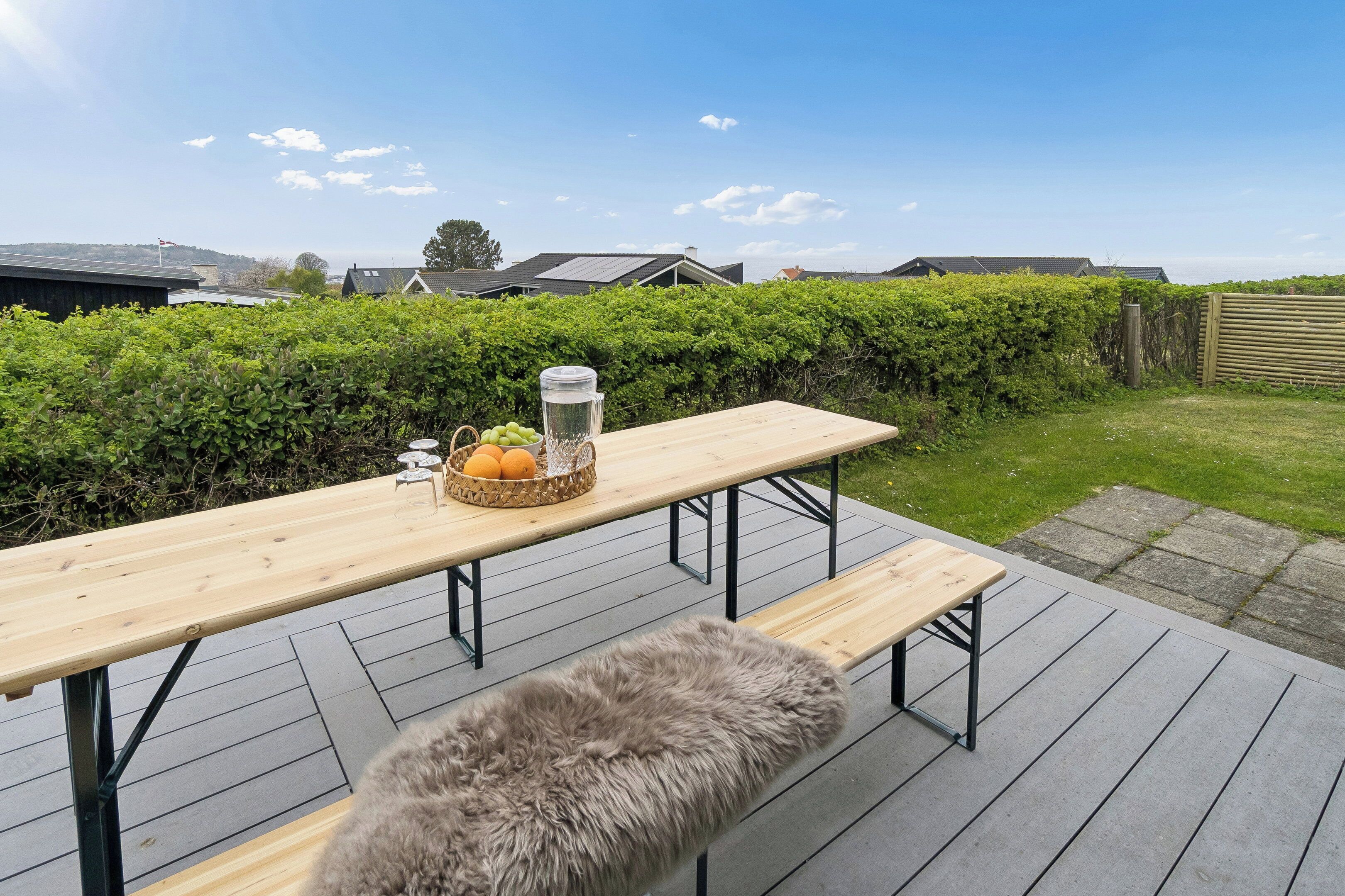 Une terrasse en bois avec un banc et une table de pique-nique, un panier rempli de fruits et un récipient d’eau.